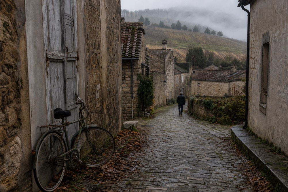 A village street in the Valpolicella hills with stone houses and vine rows behind, no text, no logos, no people