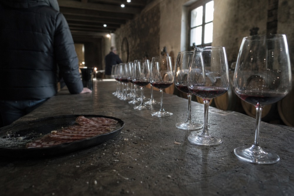 Glasses of red wine lined up on a cellar tasting counter beside a plate of salami, no text, no logos, no people