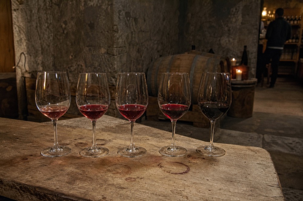 Five stemmed glasses of red wine lined on a rustic cellar tasting counter, no text, no logos, no people