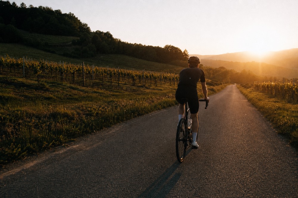 A cyclist on an e-bike on a quiet road between vineyards, with the sun low over the hills, no text, no logos, no people